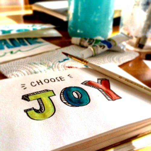 Photo of an artists pad on a desk with the words "choose joy" painted on the page