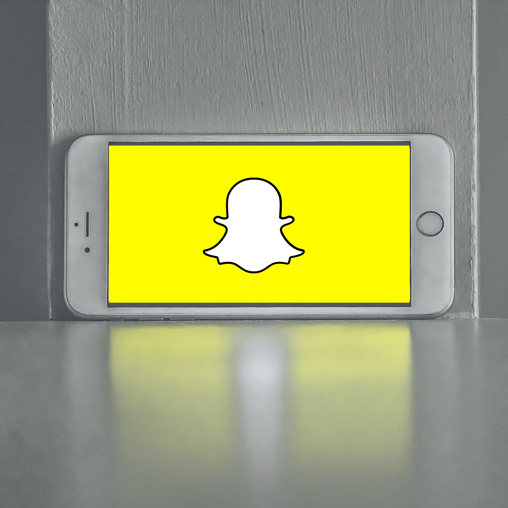 White iPhone leaning against grey concrete displaying the Snapchat logo on a yellow background. For the blog post - Snapchat Ad Specs for 2024