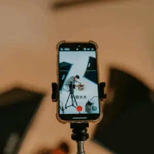 A smartphone in a tripod records a person in a studio. The setup includes lighting and a white backdrop.