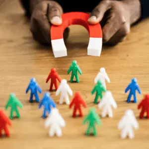 Hands hold a red U-shaped magnet attracting colorful human-shaped figures on a wooden table