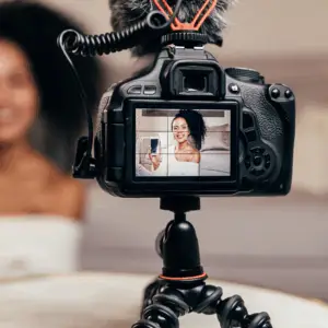 A camera on a tripod captures a smiling woman holding a smartphone.