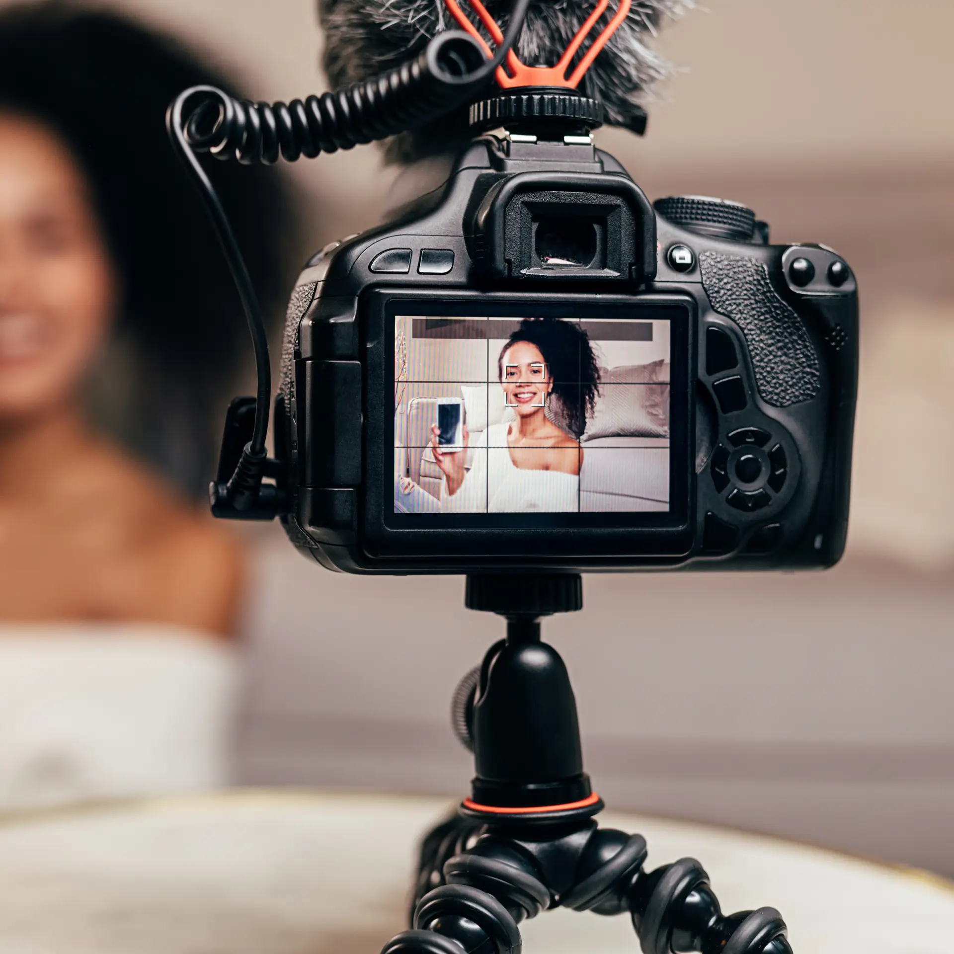 A camera on a tripod captures a smiling woman holding a smartphone.