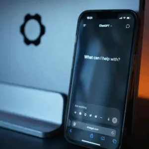Smartphone displaying ChatGPT prompt "What can I help with?" against a computer background.