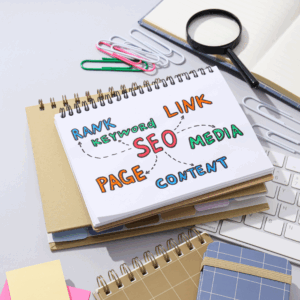 A notepad on a desk displays the word "SEO" surrounded by terms like "rank," "link," and "content." Nearby are paperclips, a magnifying glass, and notebooks.