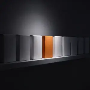 A single orange box lit by a spotlight in a row of plain white boxes on a dark shelf